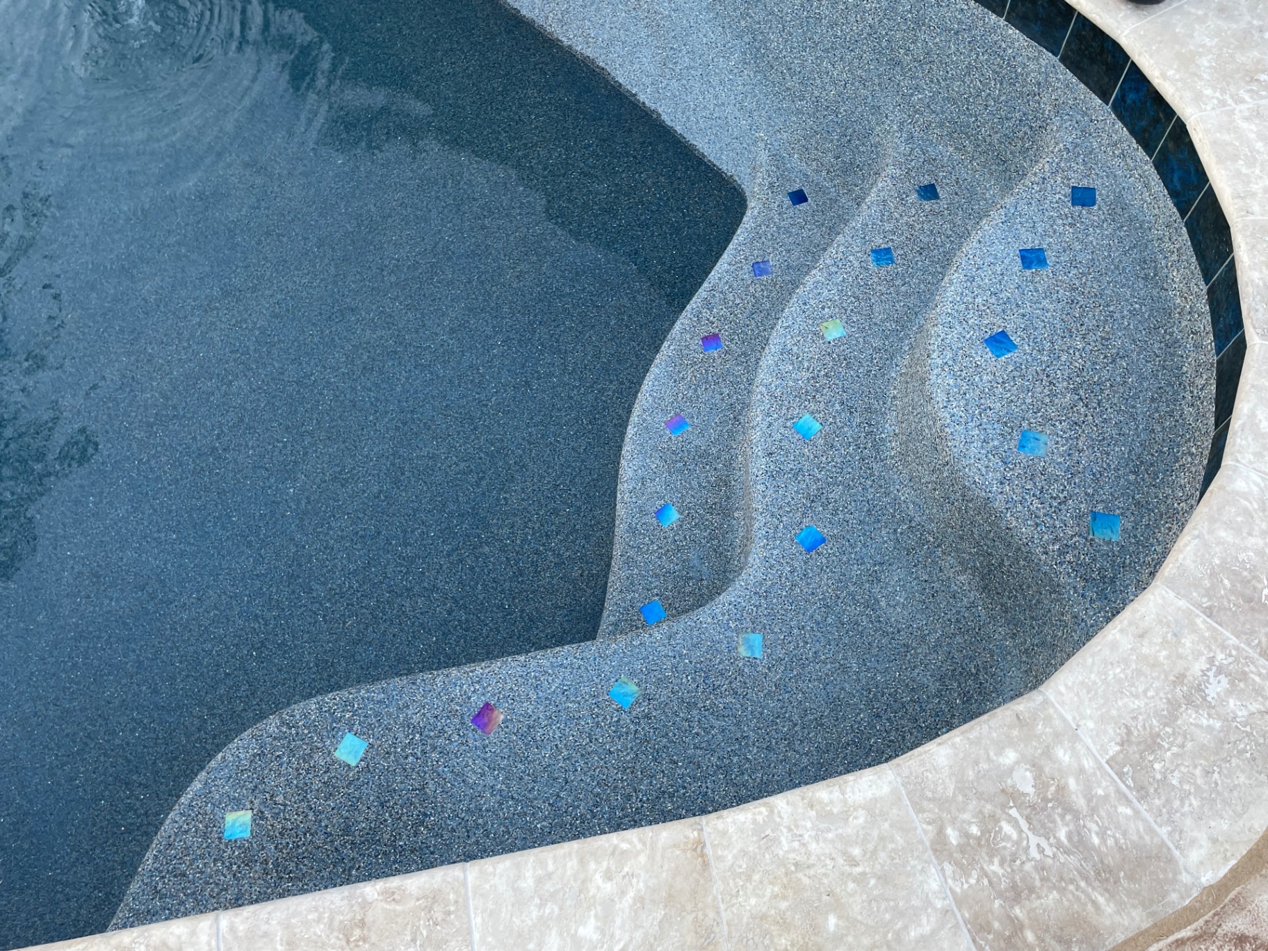 Close-up of dark pebble pool steps with iridescent blue glass tile accents