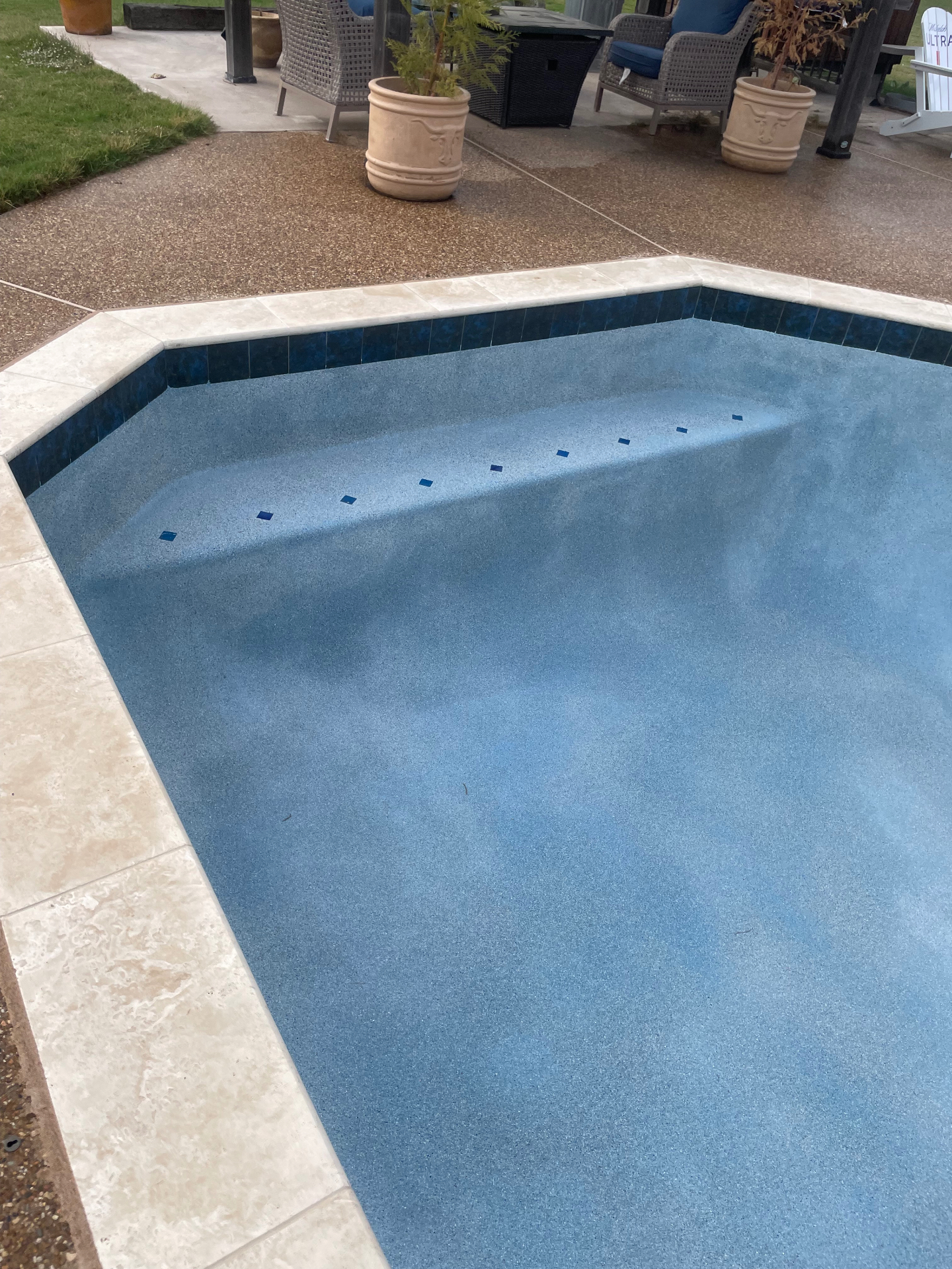Geometric pool with blue pebble finish, dark waterline tile, and travertine coping