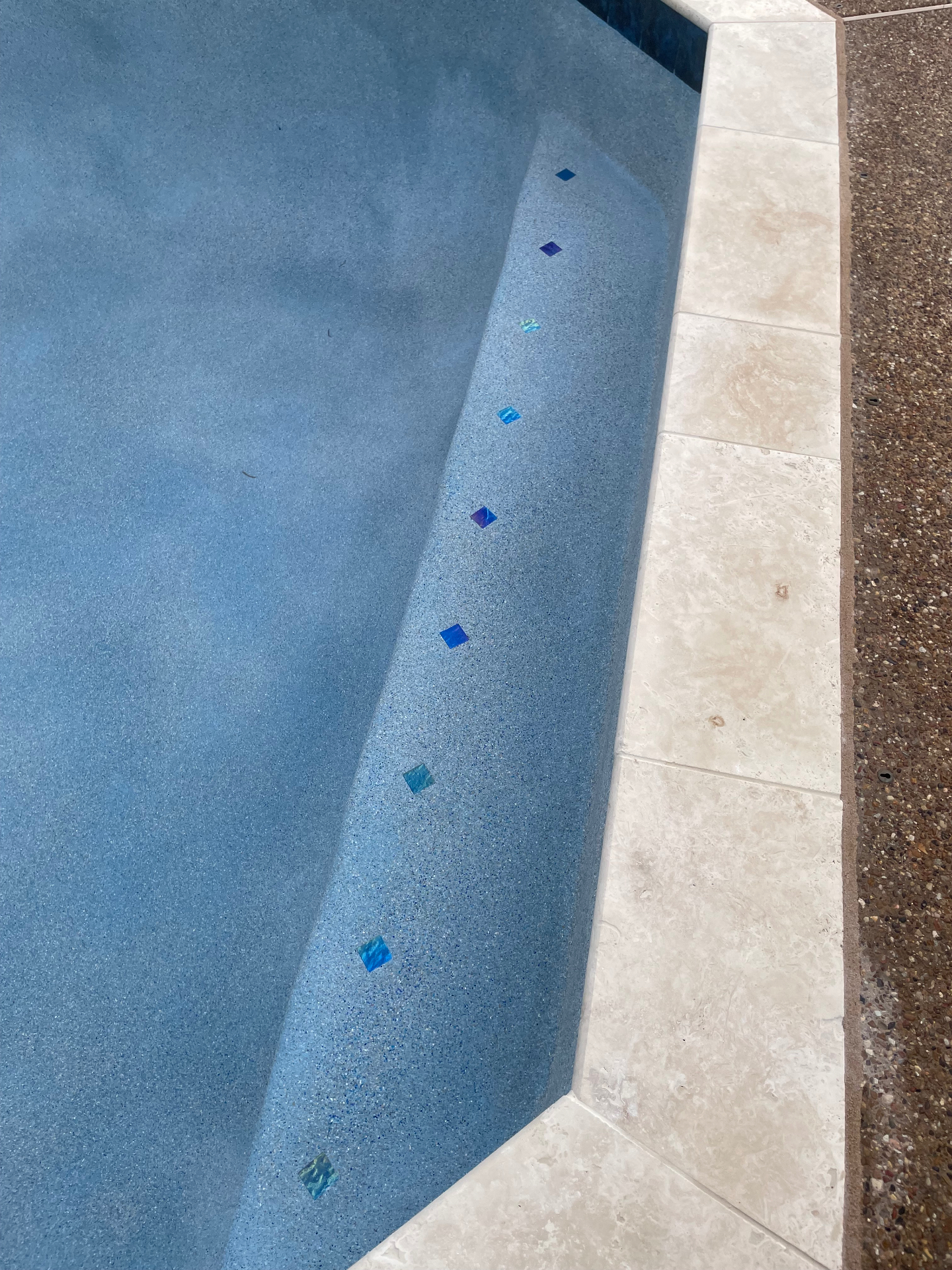 Close-up of blue pebble pool step with iridescent glass tile accents and travertine coping
